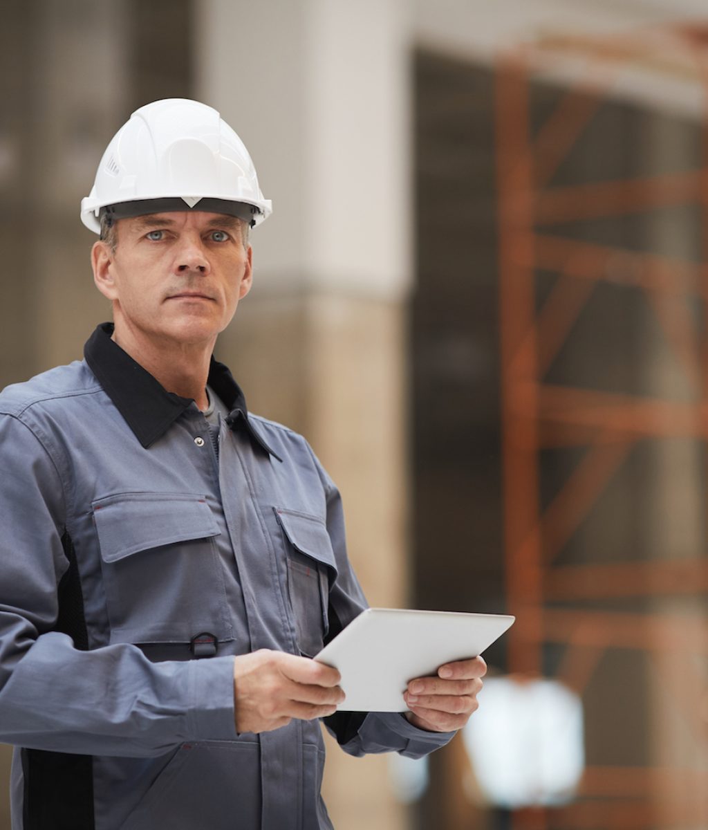 Waist up portrait of mature worker holding digital tablet and looking at camera while standing at construction site or in industrial workshop, copy space